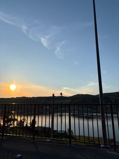 A wide-angle shot captures a sunset over a body of water, framed by a red metal railing in the foreground. The sun, a bright orb with a halo, is setting on the left side of the image, casting a warm, golden glow across the sky and water. Hills dotted with trees and houses form the distant landscape. A hot air balloon, a small red sphere, is visible in the sky to the right of the sun. A tall, dark pole stands prominently on the right side of the frame. The water reflects the sky and the trees on the shore. The scene suggests a peaceful, outdoor setting during golden hour. The location in Bartkowa-Posadowa, Poland, is not visually identifiable from the image itself, as no landmarks or specific geographical features characteristic of that location are present.