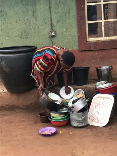 A woman in a colorful striped dress washes dishes outdoors. She sits on a large black container while cleaning pots and plates. A stack of various kitchenware including buckets, bowls, and trays is piled nearby. The scene is set against a backdrop of a weathered wall with a window and exposed electrical outlet. The ground is a reddish-brown dirt.