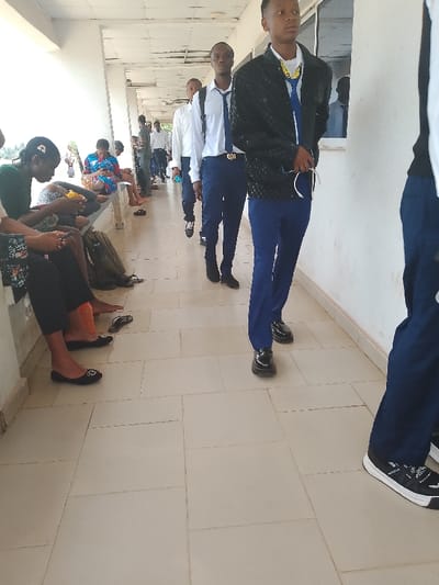 **Setting:** An indoor corridor or waiting area in Umueshi, Nigeria. The corridor is tiled and lined with a bench where people are seated. There is a window visible on the right side of the frame.

**People:**
*   A group of people are seated on a bench along the corridor wall. Some are looking at their phones.
*   A line of young men, appearing to be students, is walking in the corridor. They are wearing what seems to be a school uniform (white shirts, ties, and dark trousers). One student has a jacket on.

**Actions:** People are waiting, walking, and using their phones.

**Objects:** Benches, tiles, bags, cellphones, uniform elements (shirts, ties, trousers, shoes), a window, and a jacket.