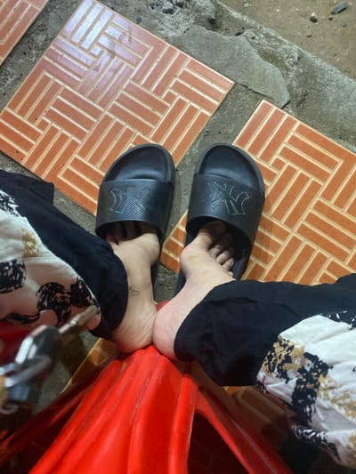 The image shows a pair of feet wearing black slides with the NY logo, resting on a red plastic chair. The feet are positioned on an outdoor tiled surface with a herringbone pattern, adjacent to a concrete edge. The person is wearing black pants and a patterned garment with black, white, and gold designs. The scene appears to be located in Watampone, Indonesia.