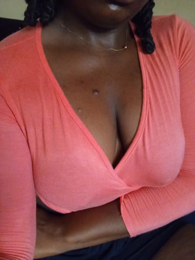 A person with dark skin is shown from the chest up, wearing a low-cut, long-sleeved coral-colored shirt and a thin gold necklace. Their hair is braided and partly visible in the top corners of the frame. Two moles are visible on their upper chest. The background is blurry and dark. The location is not identifiable in the image, and no information connects it to Bauchi, Nigeria.