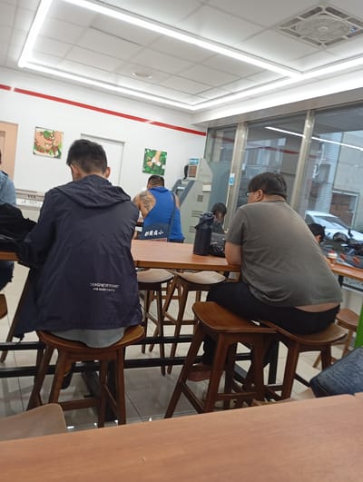 The image depicts the interior of a 7-Eleven convenience store in Kaohsiung, Taiwan, with an eating area. Two men are seated at a wooden table, facing away from the camera. The man on the left is wearing a dark blue hooded jacket with white text on the back that reads "CNSGNFNETRTFDHNRT AND SAORT FMAPLE". He is also wearing grey pants. The man on the right is wearing a grey t-shirt and black pants, with glasses and an earring. He has a visible tattoo on his back. A black thermos is on the table in front of him. Behind the men, there is a window with a view of the street and a car. The store features fluorescent lighting and a tiled floor. Several artworks are displayed on the wall.