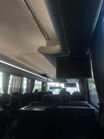 Interior shot of a bus, likely a tour bus based on the seating configuration and screen mounted in the center. The image shows rows of unoccupied seats, with visible heads and shoulders of passengers near the front and back. A small screen hangs above the middle of the bus. Curtains cover the windows. The outside view through the windows shows buildings, suggesting an urban environment. It can be seen that it is in Antwerp, Belgium because there is a bus stop sign visible through the windows that reads "Antwerpen."