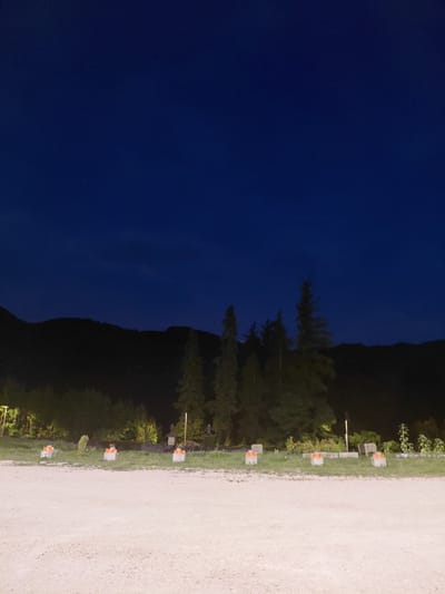 The image captures a dark, rural landscape at dusk or night. The sky is a deep, saturated blue, with faint hints of clouds. In the midground, a line of dark, silhouetted trees and hills forms a natural horizon. The foreground consists of a light-colored, gravelly ground with several concrete blocks placed at intervals. These blocks appear to have orange and yellow reflective tape on them, possibly serving as markers or barriers. The overall impression is one of a quiet, undeveloped area, possibly on the outskirts of a town. Without further context or identifiable landmarks, pinpointing the exact location within Levico Terme, Italy, is not possible.