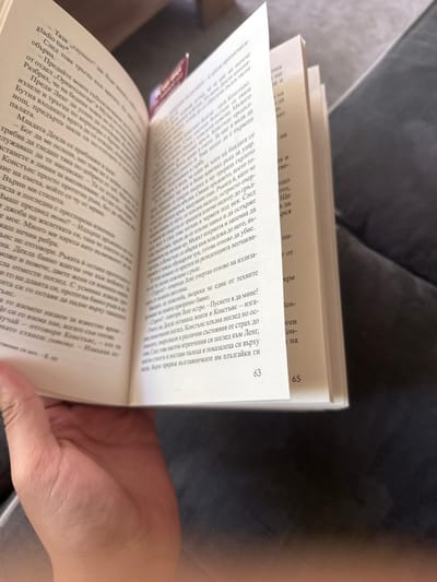 A close-up shot focuses on an open book with pages in Bulgarian, held by a fair-skinned hand. The book is on pages 63 and 65. The background is blurred, displaying a grey, textured surface.