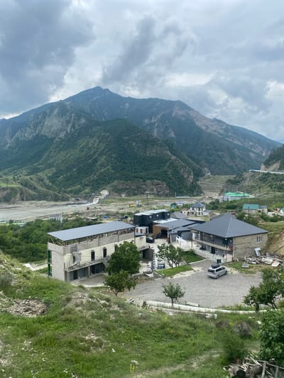The image captures a wide shot of a mountainous landscape with a village nestled in the valley. In the foreground, a partially constructed building with a grey roof and exposed concrete walls is visible, featuring a spiral staircase on its exterior. Adjacent to it is a modern-looking building with dark grey siding and a pitched roof. A gravel lot with a silver minivan parked in it is situated between these two structures.
Further into the midground, several other houses and buildings are scattered across the landscape. Some have peaked roofs, while others appear to be modular structures. A river flows through the valley, and a dam or water control structure is visible in the distance, with water cascading over it. The surrounding hills and mountains are covered in lush green vegetation, with some exposed rocky areas on the steeper slopes. The sky is overcast with heavy grey clouds. The location is identified as Chalda, Russia, an area characterized by its mountainous terrain and developing infrastructure.