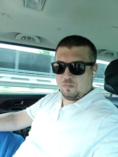 The image shows a man sitting in a car, wearing sunglasses and a white polo shirt. He has short dark hair and a beard. He appears to be looking at the camera. The car's interior is visible, including the headliner and a door handle. Outside the car window, there is a blurred view of what appears to be a highway or road. The man is wearing a white wireless earbud in his right ear. The location is identified as Ostředek, Czechia.