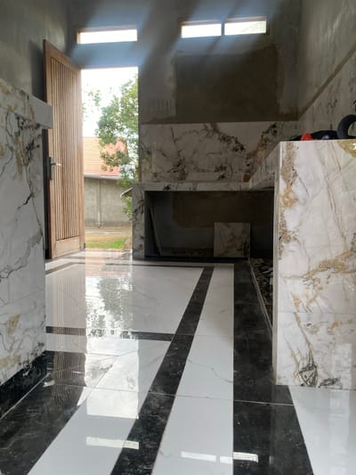 The image depicts the interior of a building under construction, likely a residential property, in Meunasahlhok, Indonesia. The focus is on the flooring and wall tiling. The floor is laid with large format tiles in a contrasting pattern of white with black and dark grey veins, creating a striking geometric design. The walls are partially tiled with marble-effect tiles, featuring white backgrounds with prominent brown and black veining, giving a luxurious appearance.
In the background, a wooden door stands ajar, revealing an outdoor view of vegetation and a red-tiled roof, characteristic of Indonesian architecture. Sunlight streams in through a high rectangular window, casting light and reflections on the polished floor. The unfinished state of the interior is evident, with exposed concrete walls in some areas and construction debris visible. There are no people or discernible actions taking place in the image, suggesting it is a captured moment during or after construction work. The overall impression is one of a modern, stylish renovation or new build, with attention given to high-contrast tiling.