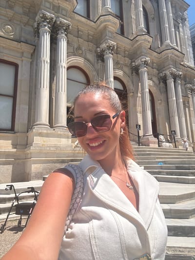 This is a vertical, medium shot photograph taken outdoors. The main subject is a young woman, positioned in the lower right quadrant of the frame and facing the camera. She has her head tilted back, smiling broadly with her teeth showing, and appears to be taking a selfie. She is wearing oversized sunglasses with dark lenses, a white denim jacket, and a delicate necklace. Her hair, a mixture of blonde and light brown, is partially visible around her face and neck.
The background features the intricate architecture of a building with ornate columns and decorative stone carvings, suggestive of a historical or neoclassical style. The building's facade is made of light-colored stone, and its texture is emphasized by the bright sunlight casting sharp shadows. To the right of the woman, a set of stairs is visible, also made of stone. In the distance, on the stairs, there appear to be a few other people and some signage, but they are too small to discern details.
The time of day appears to be daytime, likely late morning or early afternoon, given the strong sunlight and distinct shadows. The weather seems pleasant and sunny. The location is identified as Üsküdar, Turkey, which aligns with the architectural style. The overall mood of the image is cheerful and relaxed, conveyed by the woman's expression. There is no visible text in the image.