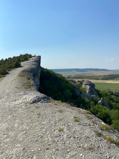 A vertical, eye-level shot captures a rocky cliff edge overlooking a vast, rolling landscape under a clear, pale blue sky. The foreground is dominated by a rough, greyish-white rocky surface covered in small pebbles and sparse, dry vegetation. To the left, a curved, worn path made of similar rocky material leads away from the viewer and curves around the edge of the cliff. This cliff edge drops sharply to the right, revealing a dense green forest that covers the slopes below. Further in the distance, the landscape opens up to reveal rolling hills, fields of green and tan, and scattered buildings, suggesting a rural or semi-rural setting. The overall impression is of a natural, elevated viewpoint offering a panoramic vista. The image appears to be taken in Kholmovka, Ukraine, as indicated by the provided context.