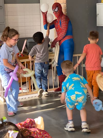 A Spiderman character in costume is interacting with children, holding two white balloons and a rope. A young girl in a light blue t-shirt and light blue pants, wearing pink sandals and glasses, is on the left side of the image, holding a purple balloon and a white balloon animal. A young boy in a grey t-shirt and blue jeans is standing behind her. Another young boy wearing a blue t-shirt with a shark print and white sneakers is in the foreground. A fourth child in an orange t-shirt and orange pants is visible to the right. The environment appears to be an indoor party space with a light gray wall, a white tiled wall, and a wooden barrier. The floor is dark gray. The image does not provide any information about the city of Mirny, Russia.