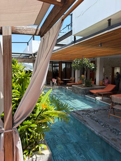 The image depicts an outdoor seating area adjacent to a swimming pool. In the foreground, a light-colored canopy is draped over wooden structures, suggesting a shaded relaxation space. Behind the canopy, lush green foliage, including tropical plants and a flowering plant with orange blossoms, thrives.
Beyond the plants, a swimming pool with a distinctive blue wavy pattern on its floor is visible. The pool area is bordered by a concrete edge with small pebbles. Further back, several lounge chairs, some with orange cushions, are arranged around the pool, alongside wooden tables and chairs. A small tree with white flowers stands in a planter near the lounge chairs.
In the background, there are glimpses of an indoor area with stone walls and wooden elements. Two individuals are partially visible inside this structure, one appearing to be carrying a box. The scene suggests a resort or a modern establishment with an emphasis on outdoor leisure and a connection to nature. The lighting indicates it is daytime, and the clear sky visible through the canopy suggests fair weather. The overall impression is one of a tranquil, upscale environment designed for relaxation. No text is visible in the image.