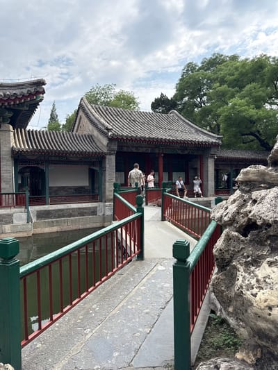 The image depicts a view of a traditional Chinese garden with a bridge over a body of water. The bridge has green posts and red railings. In the background, there are traditional Chinese buildings with tiled roofs. Several people are visible, some walking across the bridge and others sitting. To the right of the bridge, a large, textured rock formation is present. The sky is partly cloudy, and trees are visible in the background. This scene is likely located in the Xicheng District of China, known for its historical sites and imperial gardens.