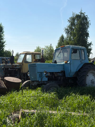 The image depicts a rural scene, possibly in Dubrova, Russia, featuring a row of old, dilapidated tractors parked near a grassy area. The tractors are of a light blue and yellow color, showing signs of rust and disrepair. Tall grass grows alongside the tractors, and trees and a clear blue sky can be seen in the background. Power lines are visible above the trees. There are no people visible in the frame.