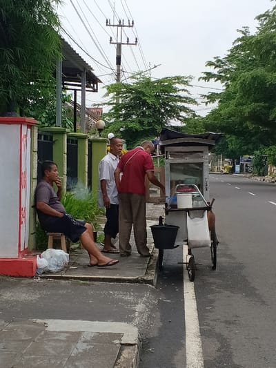 The image depicts a street scene in Ciamis, Indonesia, under an overcast sky. Three adult men are gathered near a street food cart by the side of a paved road.

On the left, a man is seated on a small wooden stool, leaning against a red and white painted wall. He wears a dark short-sleeved shirt, dark shorts, and flip-flops, and is seen smoking, with a plume of smoke rising from his hand. A white plastic bag rests on the ground beside him.

Standing next to the seated man is another man in a light-colored, possibly white, short-sleeved shirt and dark trousers, also wearing flip-flops. He is facing towards the food cart.

The third man, dressed in a red polo shirt and light brown trousers, is actively engaged with a mobile street food cart. The cart has a glass display section, wheels, and various accessories, including a black bucket hanging from the side and a white mesh bag containing what appears to be goods. He seems to be preparing food or serving.

In the background, a two-lane paved road runs alongside a sidewalk. Lush green trees line both sides of the street, and distant buildings are visible. A prominent utility pole with numerous power lines stands against the cloudy, bright sky, suggesting daytime. On the red and white wall, the red numbers "17," "18," "24," and "25" are visible. The overall scene suggests a casual, everyday street vendor and customers interaction.