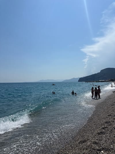 The image depicts a sunny day at a pebble beach in Finale Ligure, Italy. The turquoise waters of the Ligurian Sea gently lap at the shore, with small waves breaking and creating foam. In the foreground, the dark, textured pebbles of the beach are visible, glistening under the sun.

Several people are enjoying the water and the beach. In the middle ground of the sea, three individuals are partially submerged, with one appearing to be a child. Further out in the water, another person is visible on a yellow kayak or paddleboard.

Closer to the shore, on the right side of the image, a group of four people are standing in the shallow water, facing away from the camera. Two of them appear to be women, wearing bikinis, and they are interacting with each other. Behind them, further along the beach, more people are scattered, some swimming and others walking along the shoreline.

In the background, a rugged, dark green mountain range forms a dramatic backdrop to the scene. The sky is a clear, pale blue with a few wispy white clouds, and a bright sunbeam pierces through the atmosphere on the upper right side of the frame. The overall impression is one of a pleasant summer day at a popular coastal resort.