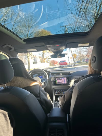From the interior of a car, a view through the sunroof reveals blue skies and bare tree branches. The driver's side interior is visible, including a black steering wheel with the Volkswagen logo, dashboard screens displaying navigation and speed, and black leather seats. Outside the car, a street scene unfolds with a red truck and surrounding houses.