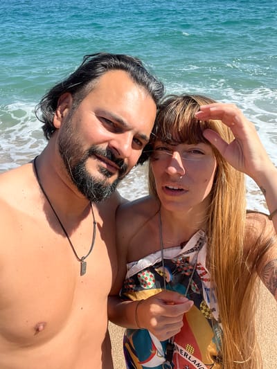 A vibrant, sunlit, close-up photograph captures a man and a woman relaxing together on a beach, with the turquoise sea and foamy waves visible in the background. The image is taken from a high angle, possibly a selfie, and is oriented so that the sea and sand run vertically alongside the subjects.

The man, positioned in the foreground, is shirtless with tanned skin, dark hair, and a full beard and mustache. He wears a simple black cord necklace with a rectangular silver pendant that clearly spells out "RAW." He looks towards the camera with a relaxed, content expression.

Next to him, or slightly above, is the woman. She has long, straight, blonde hair with bangs, and her hand gently touches her forehead or hair. She wears a colorful, patterned button-up shirt featuring fragmented text, with "WONDERLAND" partially visible, along with hints of other words. She also wears a thin necklace and a bracelet. A tattoo is partly visible on her left upper arm. Her expression is soft and appears to be smiling slightly, her mouth slightly open.

The setting is a sunny beach, with bright sunlight creating distinct shadows, indicating a clear daytime. The calm, blue-green ocean water with gentle waves creates a serene backdrop, characteristic of a coastal location such as Sant Pol de Mar, Spain. Both individuals convey a sense of peace and enjoyment.