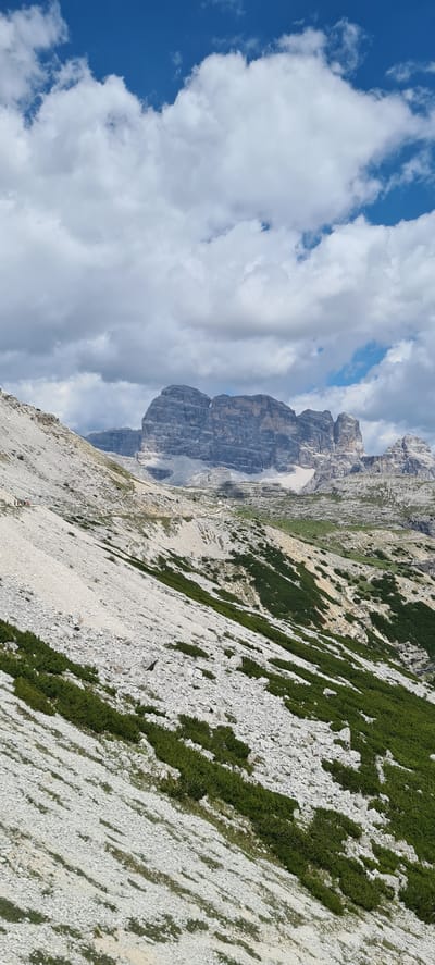 This media file captures a dramatic mountainous landscape in Auronzo di Cadore, Italy. The scene is dominated by rugged, grey rock formations characteristic of the Dolomites, with a steep, scree-covered slope in the foreground dotted with patches of vibrant green vegetation. The slope appears to be a hiking trail, with a narrow path visible winding its way up the mountainside. Two small figures, likely hikers, are discernible on this path. One is a distant red figure, and the other, a dark silhouette, is further up the slope.

The sky is a brilliant blue, interspersed with large, fluffy white clouds that cast shadows on the mountains. The lighting suggests it is daytime, likely mid-morning or early afternoon, given the bright sunlight and clear visibility. The terrain is challenging and wild, conveying a sense of adventure and the raw beauty of alpine environments. The perspective is from a high vantage point, looking across and down the steep mountainside towards more distant peaks shrouded in haze. No text is visible in the image.