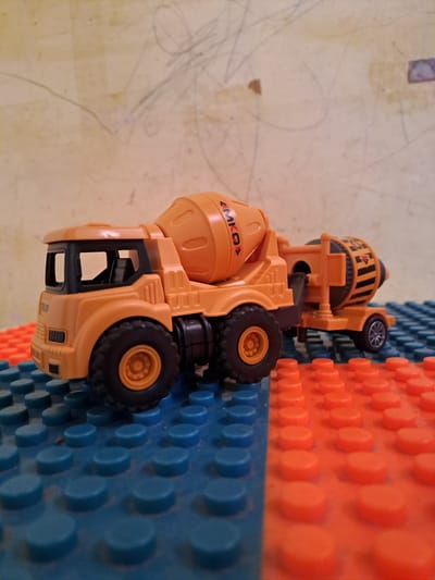 A close-up, eye-level shot depicts an orange toy cement mixer truck resting on a surface made of blue and orange plastic building blocks. The background is a light-colored wall with faint scribbled markings. The truck is positioned diagonally across the frame, with its front end on the left and its rear extending towards the right. The truck's main drum is tilted, and the cab is visible with its windows. The building blocks are arranged in a grid pattern. The lighting appears to be coming from the front and slightly to the left. There are no people visible in the image.