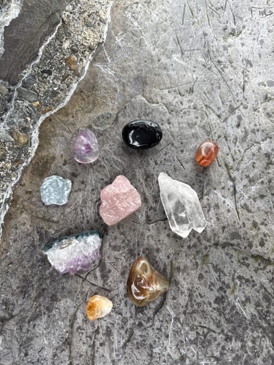 The image shows a collection of polished and raw gemstone specimens arranged on a cracked, grey stone surface. The specimens include a red-orange carnelian, a clear quartz crystal, a black onyx, a pink rose quartz, a translucent brown agate, a light yellow citrine, and several amethyst pieces with varying degrees of crystal structure and color saturation. The stone surface appears to be a large, fractured rock, possibly located in an outdoor environment. The location, Llanberis, United Kingdom, is only based on the user request. No people, interactions, or actions are visible. The environment appears to be an outdoor setting given the natural stone and lighting.