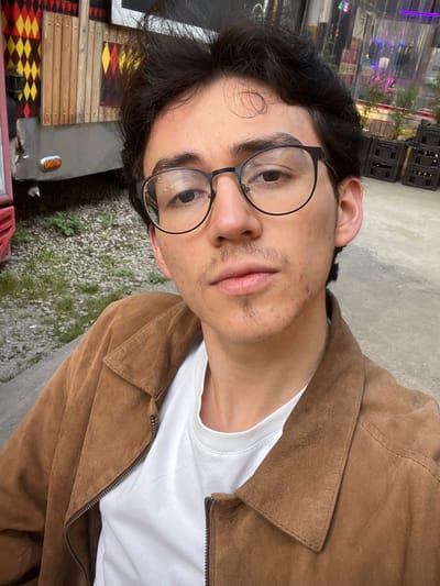 A male subject, appearing to be in his late teens or early twenties, is depicted in a medium close-up, centered and facing directly forward. He has dark, wavy hair, wears black-framed glasses, a thin mustache, and a small soul patch. Visible skin blemishes are present on his chin and right cheek. He is dressed in a white crew-neck t-shirt and an unzipped brown or tan suede-like jacket.

The scene is an outdoor commercial or recreational area, likely a patio or open-air market, located in Bogota, Capital District, Colombia. To the subject's left, a portion of a converted vehicle or structure, resembling a food truck, is visible. It features a grey metallic lower section with a horizontal red stripe and an orange reflector light. Above this, wooden paneling displays a red, yellow, and black diamond pattern. The ground in this section is unpaved, consisting of gravel or dirt with sparse green vegetation. To the subject's right, a transparent plastic or tarp barrier encloses an area containing several dark, stacked crates and blurred green foliage. Faint purple and white ambient lighting is discernible through the barrier. The immediate foreground and the ground surrounding the subject consist of a light-colored concrete or paved surface. The subject is stationary, looking directly at the camera.