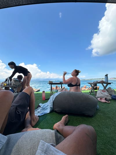A group of people relax on what appears to be a boat or a deck next to the ocean. One woman, dressed in a black bikini, is drinking from a glass. Another person in a black outfit is bending over, possibly stretching. Pillows and blankets are scattered around, and some individuals are lounging in the background. A small wooden table holds drinks and personal items. The sky is blue with fluffy white clouds.