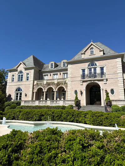 This media file depicts a grand, multi-story building, likely a mansion, set against a clear, deep blue sky. The building showcases elaborate architecture, primarily constructed from light-colored brick with extensive stone detailing, including cornices, window frames, and balustrades. It features multiple gabled roofs with dark shingles, and a variety of windows ranging from large arched and semicircular designs to rectangular panes, some adorned with decorative wrought iron balconies.
A prominent arched entryway with dark double doors is accessible via a set of stairs, flanked by manicured topiary shrubs and decorative urns. Stone balustrades line various terraces and balconies, some supported by arched columns.
In the foreground and running parallel to the building, a long, multi-tiered stone fountain or water feature is visible, with water gently flowing and creating small jets. Lush, well-maintained green hedges line both sides of the fountain and the path leading to the building's entrance. Some patches of the hedges show slight browning.
The scene is bathed in bright sunlight, indicative of a clear mid-morning or early afternoon, with distinct shadows cast by the architectural elements and foliage. No people or visible text are present in the image. The opulent architecture and meticulously maintained landscaping suggest a high-end residential estate in an affluent area.