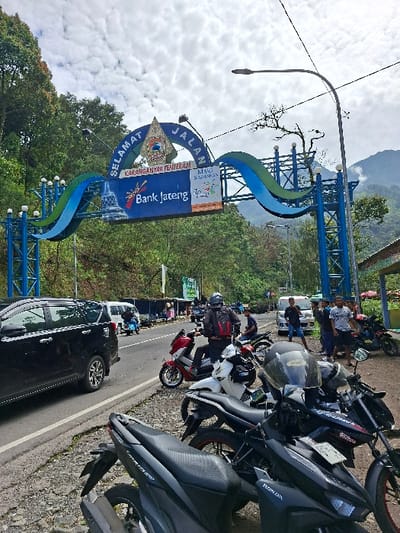 The image shows a scene on a paved road with a blue archway sign that reads “Selamat Jalan” (Welcome).  The archway also has a bank sign that reads “Bank Jateng” (Bank Central Java). The sign is located in Kecamatan Tawangmangu, Indonesia, at the coordinates -7.663, 111.188.  The image is taken from a perspective standing near the bottom of a hill and looking up the road towards the archway, showing four motorcycles parked on the side of the road and several people standing near the motorcycles. On the left side of the road, a black car is parked in a parking lot. The road continues on beyond the archway, going into a forest with hills in the background. There is a cloudy sky with a few white clouds.