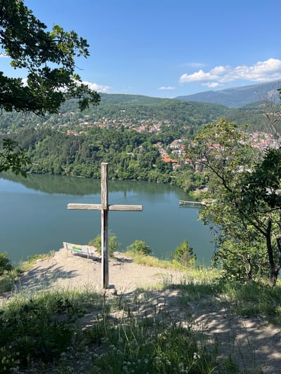 A wooden cross stands on a rocky outcrop overlooking a wide, placid lake. The lake's surface reflects the lush green trees that line its shores and the scattered houses of a town nestled in the rolling hills beyond. A blue sky with scattered white clouds completes the picturesque scene, with the foreground showing dry grass and small wildflowers. In the background, a pier juts out into the water, and a faint structure is visible on the left side of the lake. A park bench with a colorful informational sign is positioned near the cross, offering a place to rest and admire the view. This vantage point is a popular spot in Pancharevo, Bulgaria, known for its scenic beauty and tranquil atmosphere.