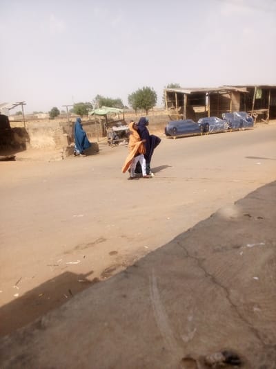 This image captures a street scene in Tudun Murtala, Nigeria, under bright, possibly midday, lighting conditions. The foreground consists of an unpaved road or dirt-covered asphalt surface, with a low concrete barrier or curb on the right.
Three individuals are visible. On the left side of the road, a person wearing a full-length blue garment stands facing left, positioned near a dirt mound and a rudimentary structure. In the center-right of the road, two individuals are in close proximity, appearing to walk or stand together. One person wears an orange or light brown full-length garment over white trousers, while the other wears dark blue or black full-length attire over what appears to be green trousers.
The environment includes several structures. On the immediate right, a series of open-fronted, wooden stalls with corrugated metal roofs line the road. In front of these stalls, several pieces of blue upholstered furniture, possibly sofas or chairs, are visible, individually wrapped in clear plastic and resting on wooden pallets. Further in the background, more distant buildings and scattered green trees are visible under a hazy sky. Debris, including small pieces of litter and dark stains, is present on the road surface.