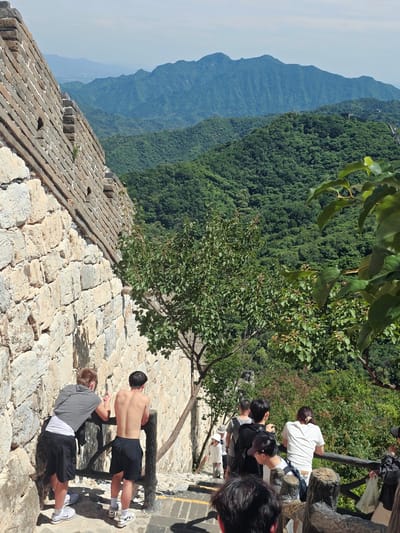 This image depicts a group of people at the Great Wall of China in Huairou District, China. The scene is set on a bright, sunny day with clear skies, suggesting it's mid-morning or afternoon. The Great Wall stretches across a mountainous, densely forested landscape, with layers of hazy blue mountains receding into the distance. The wall itself is constructed from weathered stone and brick, with a wooden railing in the foreground.
In the foreground, two young men are engaged in physical activity. One, shirtless, is performing push-ups with his hands on the wooden railing, while the other, wearing a grey shirt and black shorts, is in a plank position on the stone ground. Both are wearing athletic shoes.
Further back, several other individuals are visible, some looking out at the scenic vista, others walking along the wall. One person in a white shirt is leaning on the railing, observing the view. The people appear to be tourists, enjoying the historical site and its natural beauty. The overall atmosphere is one of leisure and exploration. No visible text is present in the image.