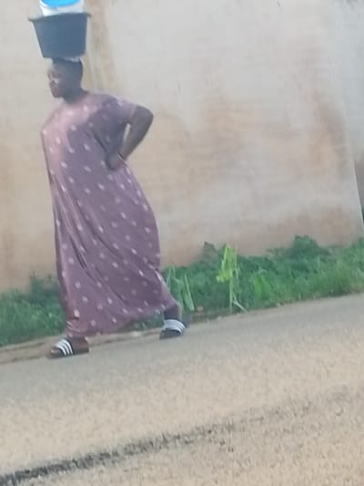 A person walks along a paved surface, carrying a dark bucket with a blue lid balanced on their head. The person is wearing a long, mauve dress adorned with white polka dots and white and black striped sandals. They are walking on a paved road with some green vegetation and a light-colored wall in the background. The scene appears to be in Rafin Bauna, Nigeria.