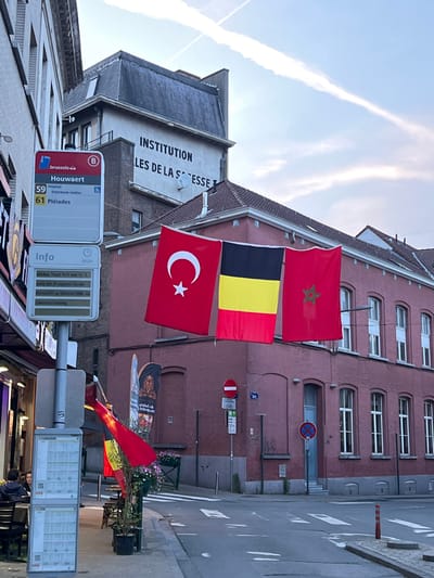 The image captures a street scene in Saint-Josse-ten-Noode - Sint-Joost-ten-Node, Belgium, with flags of Turkey, Belgium, and Morocco prominently displayed, hung from the facade of a red brick building. In the foreground, a bus stop sign for "Houwaert" with route numbers 59 and 61 is visible. The bus stop displays information in a language consistent with French or Dutch, suggesting it's a public transport point within the Brussels-Capital Region. Behind the bus stop, a building with "INSTITUTION LES DE LA S ESSE" is inscribed on its facade. The environment suggests an urban setting with a paved street, sidewalks, and other buildings. The sky has contrails from aircraft, indicating daytime. A few individuals are seated at tables outside a building to the left, appearing to be patrons of a cafe or restaurant. Several traffic signs, including parking restrictions, are also present.