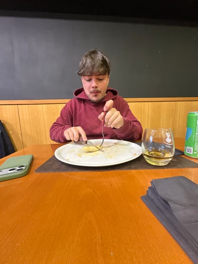 A male subject with short, dark brown hair and light stubble is seated at a light-colored wooden table in an indoor restaurant setting located in Peso da Régua, Portugal. He wears a dark red hooded sweatshirt and is actively using a knife in his right hand and a fork in his left hand to cut a small, light-yellow piece of food on a white, mostly cleared plate. The plate rests on a dark rectangular placemat. To the subject's left, a light green-cased smartphone with multiple camera lenses is positioned on the table. To his right, a stemless glass filled with an amber liquid, bearing a "1825" logo, and a green Sprite soda can are visible on the same dark placemat. A stack of black paper napkins is also present on the table to his right. The background features light wooden paneling across the lower section of the wall, topped by a plain, dark gray wall.