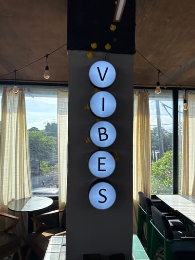 Captured from an unconventional angle, the image presents a room scene, likely from a commercial establishment, situated in Uluwatu, Indonesia. The focal point is a sign composed of five circular lights, each illuminating a letter to spell out "VIBES". Small yellow rubber ducks are visible atop the lights. The sign is affixed to a grey structure near windows. Through the windows, greenery and buildings typical of an urban environment are visible. Translucent curtains adorn the windows, softening the natural light. String lights with Edison-style bulbs stretch across the ceiling. The presence of furniture, including tables and chairs, suggests a cafe or similar establishment. The image is taken from a low angle, giving a unique perspective of the scene.