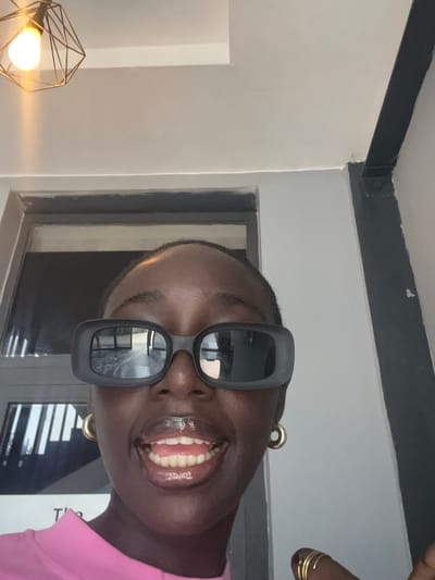 This is a close-up, eye-level, selfie-style photo of a young Black person wearing large, rectangular, black-framed sunglasses and a bright pink top. The person is smiling broadly, with their mouth open, showing their teeth, and appears to be looking directly at the camera. They have gold hoop earrings in their ears and gold rings on one hand. The background is dimly lit, with a light-colored wall and a dark beam running horizontally across the top of the frame. There's a window with blinds visible behind the person, reflecting some of the interior. In the lower left corner, part of a modern, geometric lamp with a glowing light bulb is visible. The overall impression is casual and joyful. The image is oriented vertically. The scene appears to be indoors, possibly in a home or a well-lit lounge area. The lighting suggests it could be daytime, but the indoor setting makes it difficult to ascertain. There is no visible text in the image.