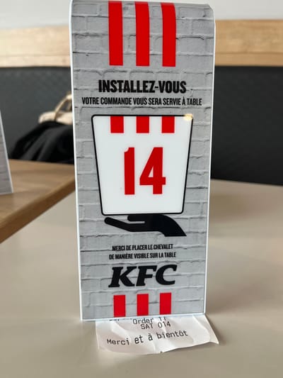 The image shows a table tent at a KFC restaurant. The tent is printed with a faux brick wall pattern and features the KFC logo, the number "14," and the phrase "INSTALLEZ-VOUS VOTRE COMMANDE VOUS SERA SERVIE À TABLE," which translates to "Make yourself comfortable, your order will be served at the table." It also includes the text "MERCI DE PLACER LE CHEVALET DE MANIÈRE VISIBLE SUR LA TABLE," which translates to "Please place the tent visibly on the table." A receipt is protruding from the side of the tent, with the order number "014" and the phrase "Merci et à bientôt," which translates to "Thank you and see you soon." The restaurant is located in Andrézieux-Bouthéon, France.