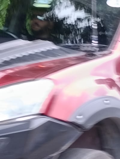 This is a close-up, low-angle, motion-blurred photograph taken from the side of a red vehicle, likely a truck or SUV, in Manali, India. The vehicle appears to have a dent or damage on its side, near the headlight. The reflection on the vehicle's surface shows a blurry image of greenery and a person's face. The image is highly abstract due to motion blur and focus issues, making it difficult to discern specific details of the surroundings or the person. The environment appears to be outdoors, possibly on a street or road, with the presence of foliage.
