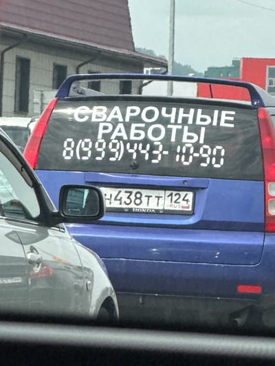 The image shows the rear of a blue car parked on a street. On the rear window, there is a black sticker with white text that reads "СВАРОЧНЫЕ РАБОТЫ 8(999)443-10-90". This translates to "Welding Services 8(999)443-10-90". A license plate is also visible on the rear of the car, reading "438TT 124 RUS". The scene appears to be in a residential area, with a building with a red roof and white walls visible in the background. The sky is overcast, suggesting a cloudy day. The overall impression is of a utilitarian vehicle advertising a service. There are no people visible in the image.