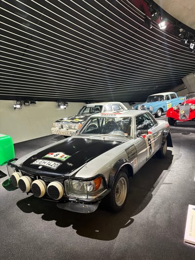 In a museum exhibit in Stuttgart, Germany, a collection of vintage Mercedes-Benz cars is displayed on a dark, carpeted floor. The cars are illuminated by spotlights from above, casting long shadows. In the foreground, a silver Mercedes-Benz rally car with a black hood and racing decals is prominently featured. It has a license plate that reads "S-EH 1420". To its left, a portion of a bright green object is visible. Behind this car, another Mercedes-Benz sedan, also silver with a black hood and racing decals, is parked. Further back, a light blue Mercedes-Benz sedan and a bright red vintage Mercedes-Benz sports car are visible. The ceiling of the museum is characterized by black panels with thin white stripes, creating a striped pattern. A sign is visible on the floor to the right of the red car, which appears to be a display plaque.