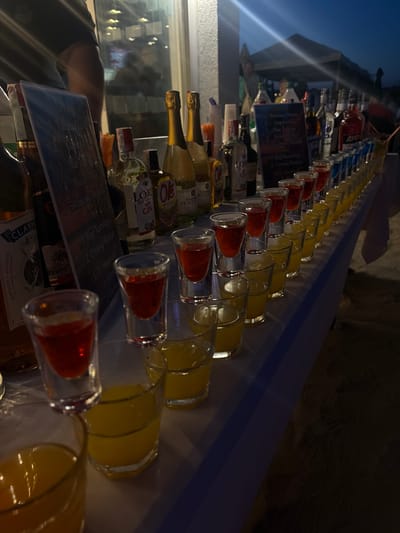 The image shows a long table laden with rows of shot glasses filled with various colored liquors. The table is set up outdoors, likely at a bar or event venue, with a dimly lit background suggesting it is dusk or nighttime. In the foreground, several shot glasses are filled with a red liquid, followed by rows of yellow and blue liquids. Behind the shot glasses, there are numerous bottles of alcohol, including champagne, gin, and other spirits. A menu board listing cocktails is visible on the left side of the image. The scene appears to be in Kranevo, Bulgaria, possibly at a beachside bar given the presence of sand and an outdoor tent in the background. A person's arm, wearing a dark t-shirt, is visible on the far left, suggesting someone is tending to the bar. The overall ambiance is festive, with the array of colorful drinks and bottles hinting at a party or gathering.