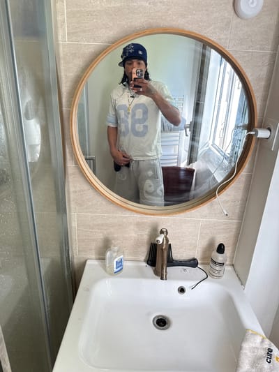 A full-body mirror selfie taken in a bathroom shows a person with dark hair, wearing a blue baseball cap, a light-colored jersey with the number "88" in blue, and light-colored shorts. The person is holding a phone in their right hand, taking the photo. The bathroom features light-colored tiled walls, a white sink with a faucet, a bottle of soap, a comb, and a bottle of nasal rinse. A shower enclosure is visible on the left side of the image, and a window is reflected in the mirror. The scene appears to be a personal bathroom in Massy, France.