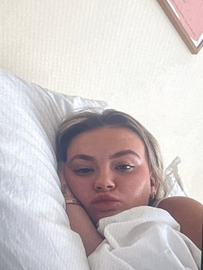 A close-up, high-angle shot shows a young Caucasian woman lying in bed. She is on her left side, with her head resting on a white pillow. Her face is turned towards the camera, and she is looking directly at it with a neutral expression. Her blonde hair is messy, and she is wearing a gold ring on her right hand. The background is a plain white wall with a textured pattern. The room appears to be a bedroom. The image is taken with a mobile phone and appears to be a selfie. The lighting is natural and soft, suggesting it was taken during the day. There are no visible indications of the location within the city of Pruszcz Gdański, Poland, other than what can be inferred from the general setting of a domestic bedroom.