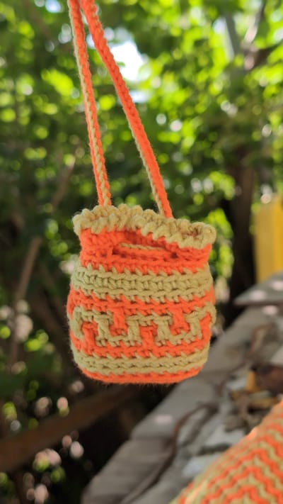 An orange and tan crocheted bag with a patterned design hangs from two crocheted straps. The bag is small and cylindrical, with a ruffled top edge. The background is blurred and consists of green foliage, suggesting an outdoor setting. The image does not provide any information about the city of Kostiantynivka, Ukraine, or any specific location within it.