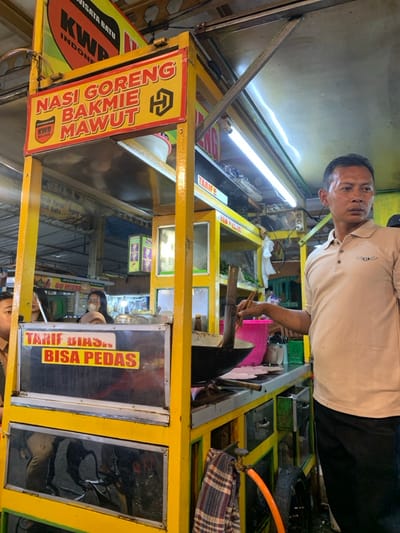 The image captures a detailed view of a food stall operating indoors within a market or food court in Kota Batu, Indonesia. The central subject is a bright yellow food cart, prominently featuring a top sign with red and black lettering that reads "Nasi Goreng Bakmie Mawut" and "KWB Wisata Batu Indonesia," confirming the location. A male vendor, with short dark hair and a beige polo shirt bearing a dark emblem, stands on the right side of the cart, facing left. He holds cooking utensils within a dark wok positioned on the cart's cooking surface. To the left and behind the cart, a female individual wearing a light-colored face mask is partially visible. Another person's head and shoulder are partially visible on the far left. A mid-level sign on the cart's front panel displays "Tarif Biasa. Bisa Pedas." (Normal price. Can be spicy.) in red and yellow text on a metallic background. Ancillary objects on the cart include a pink plastic bucket, green containers, and an orange hose connected to a valve near the bottom, with a plaid cloth hanging nearby. The environment is illuminated by overhead fluorescent tube lights, and a metal roof structure is visible. Blurred background elements suggest other market stalls or activity.