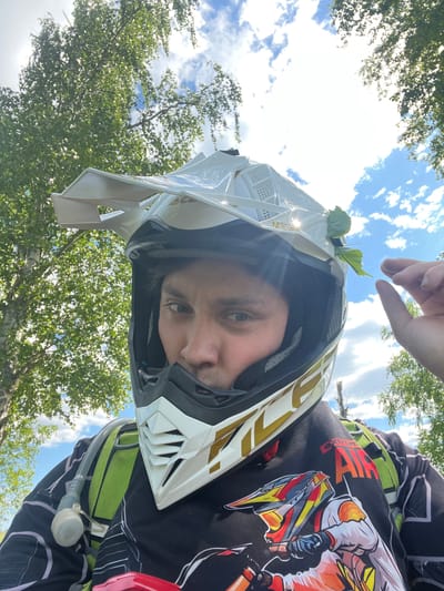 This low-angle, close-up outdoor portrait, taken in Новочувашский, Russia, captures an individual from the chest up, wearing a white and gold motocross helmet (branded "Acerbis") and a black long-sleeved jersey. The subject, likely male, looks directly into the camera; his face is partially obscured by the helmet, revealing his eyes, nose, and mouth, with dark hair visible underneath. A small green leaf is visibly tucked into the right side of the helmet, near the visor. A green-strapped hydration pack is visible over the left shoulder, with a clear plastic drinking tube and bite valve clipped to the front strap. The black jersey features a colorful graphic of a dirt bike rider and the text "Xtreme AIR" in red and white on the upper left chest. The background consists of a bright blue sky with scattered white cumulus clouds, framed by lush green foliage of deciduous trees, consistent with birch, particularly in the upper left. Strong sunlight illuminates the scene, casting highlights on the helmet and leaves. A hand, likely the subject's right, is partially visible in the upper right quadrant of the image, with fingers extended.