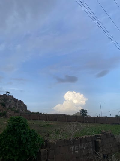 In the city of Barikin Jirga Summa, Nigeria, a natural landscape unfolds under a partly cloudy sky. In the foreground, a rough brick wall, partially constructed and bearing some graffiti, marks the edge of a plot of land. Beyond this, a dense cluster of green foliage dominates the left side of the frame, reaching upwards. Further back, a sturdy, brick wall stretches horizontally across the midground, delineating a property. Beyond this wall, a grassy expanse dotted with sparse vegetation extends towards a rocky outcrop on the left. This outcrop is steep and weathered, with a lone tree perched precariously near its summit. The sky above is a canvas of blues and whites, with several clouds scattered across it, including a prominent, fluffy white cumulus cloud that stands out against the lighter shades. Electrical wires crisscross the upper portion of the image, suggesting the presence of infrastructure. The scene appears to be an undeveloped or semi-developed area, characterized by natural rock formations and rudimentary construction.
