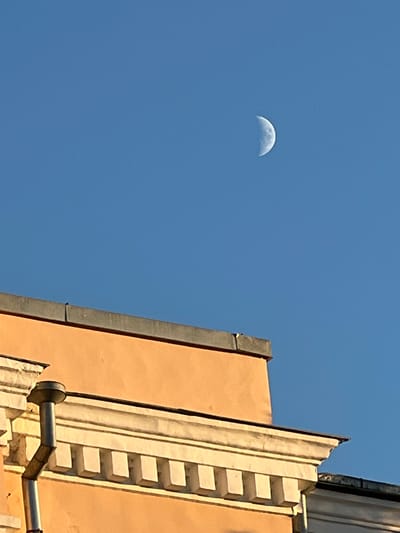 The image depicts a clear blue sky with a crescent moon visible in the upper right quadrant. In the foreground, the upper section of a building is shown. The building has a peach-colored facade with decorative white architectural elements, including a cornice with dentils. A downspout is attached to the left side of the building. The scene is illuminated by sunlight, casting a warm glow on the building. The location is Tver, Russia.