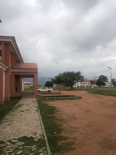 The image depicts an outdoor scene at the Federal University, Wukari, Nigeria, under an overcast sky. In the foreground, a light-colored brick walkway, partially overgrown with grass, leads towards a building. To the left, a portion of a salmon-colored building with white trim is visible, featuring prominent white columns and a red roof. A person is seated in the covered entrance of this building. Further into the scene, a white pickup truck is parked on a dirt and grass area. Beyond the truck, several buildings with white facades and red roofs are visible in the distance, interspersed with trees. Solar-powered streetlights are also present in the middle ground. The ground consists of reddish-brown dirt and patches of grass. The overall impression is that of a campus environment on a cloudy day.